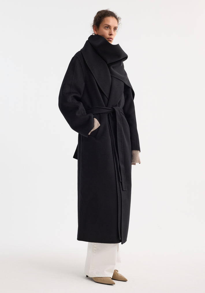 Signature double-faced scarf coat with belt in black, worn by a woman. Features oversized collar, belted waist, side pockets; styled with white trousers, nude heels.