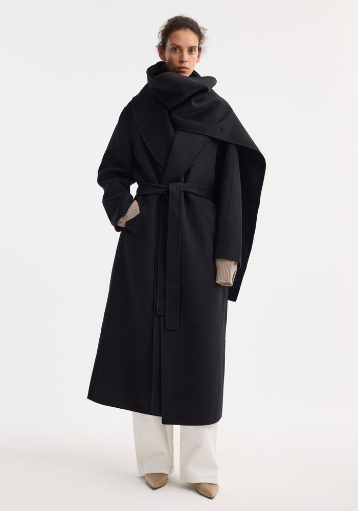 Model wearing the Signature double-faced scarf coat with belt, showcasing its sculptural scarf, wrap style, and taupe fingerless gloves, paired with white trousers and beige heels.