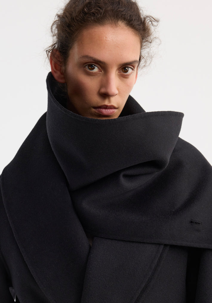 Signature double-faced scarf coat in black, showcasing its voluminous wool fabric draping elegantly around the model’s shoulders, with visible wrap belt and side pockets.