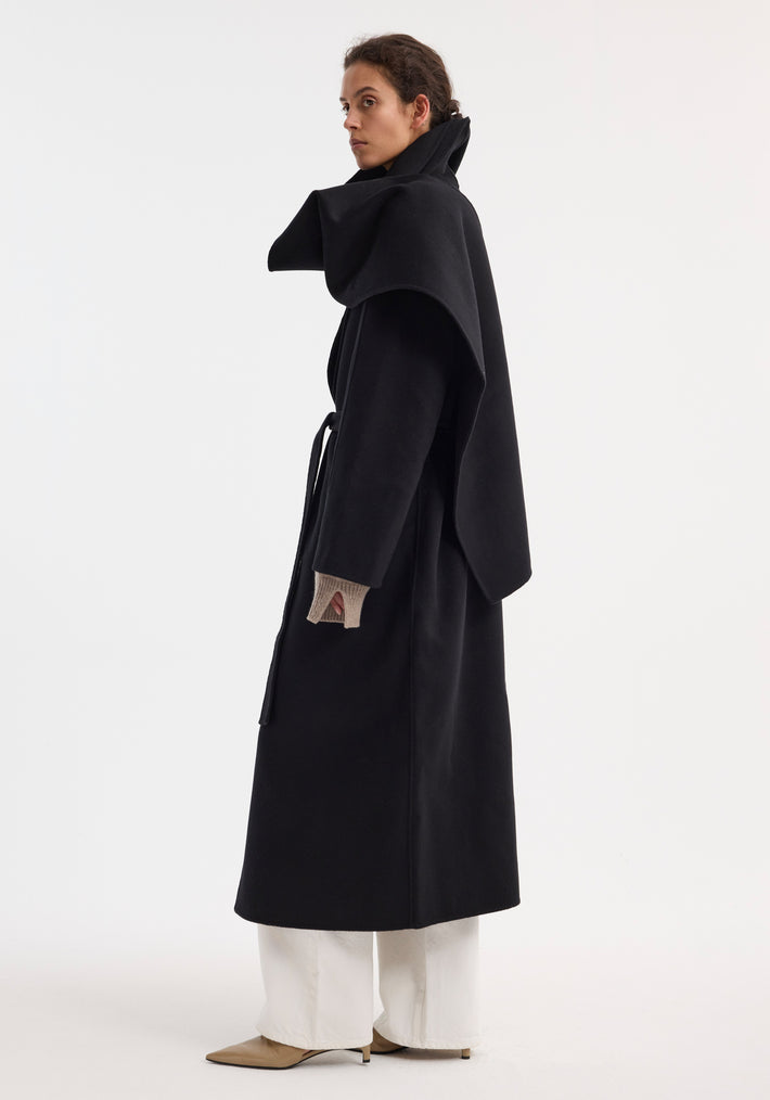 Signature double-faced scarf coat with belt, featuring an oversized draped collar, wrap belt, side pockets, worn with tan fingerless gloves and white trousers.