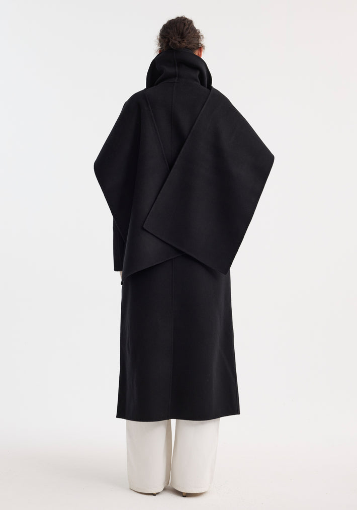 Signature double-faced scarf coat with belt, featuring a high funnel neck and wrap-style shoulder panel, worn with loose white trousers. Model's hair in a bun.