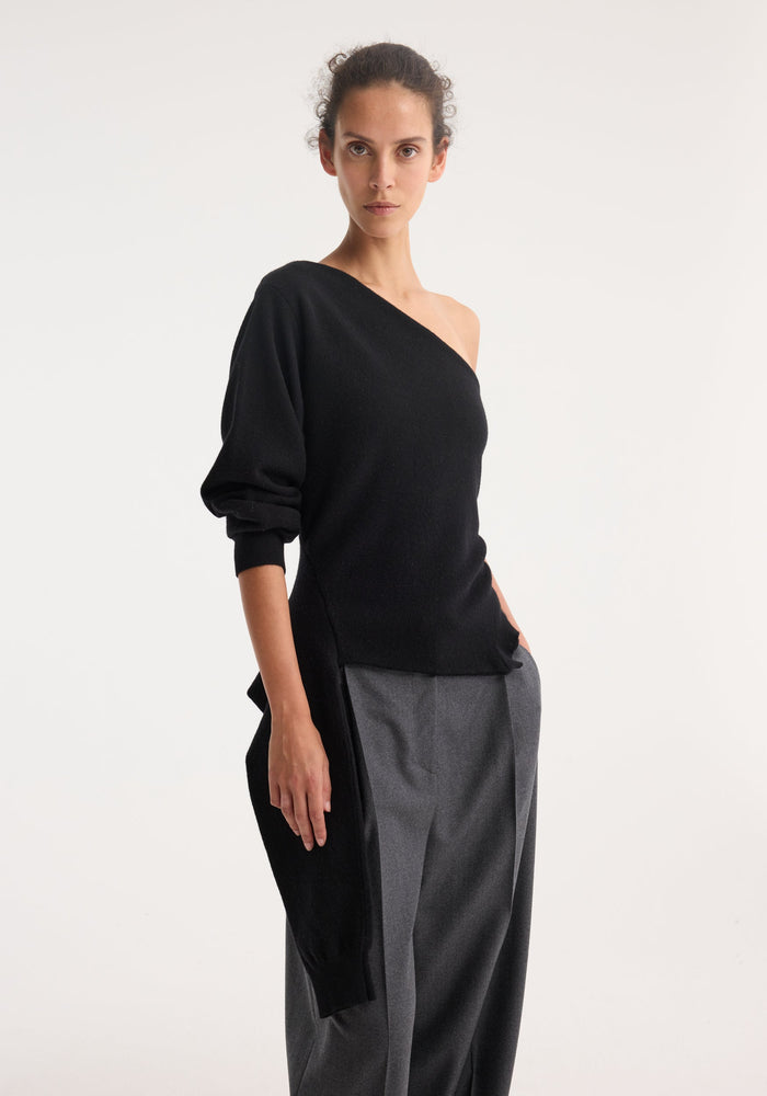 Model wearing Off shoulder single sleeve top in black, showcasing its asymmetric wool-cashmere design with draped hemline, paired with high-waisted grey trousers.