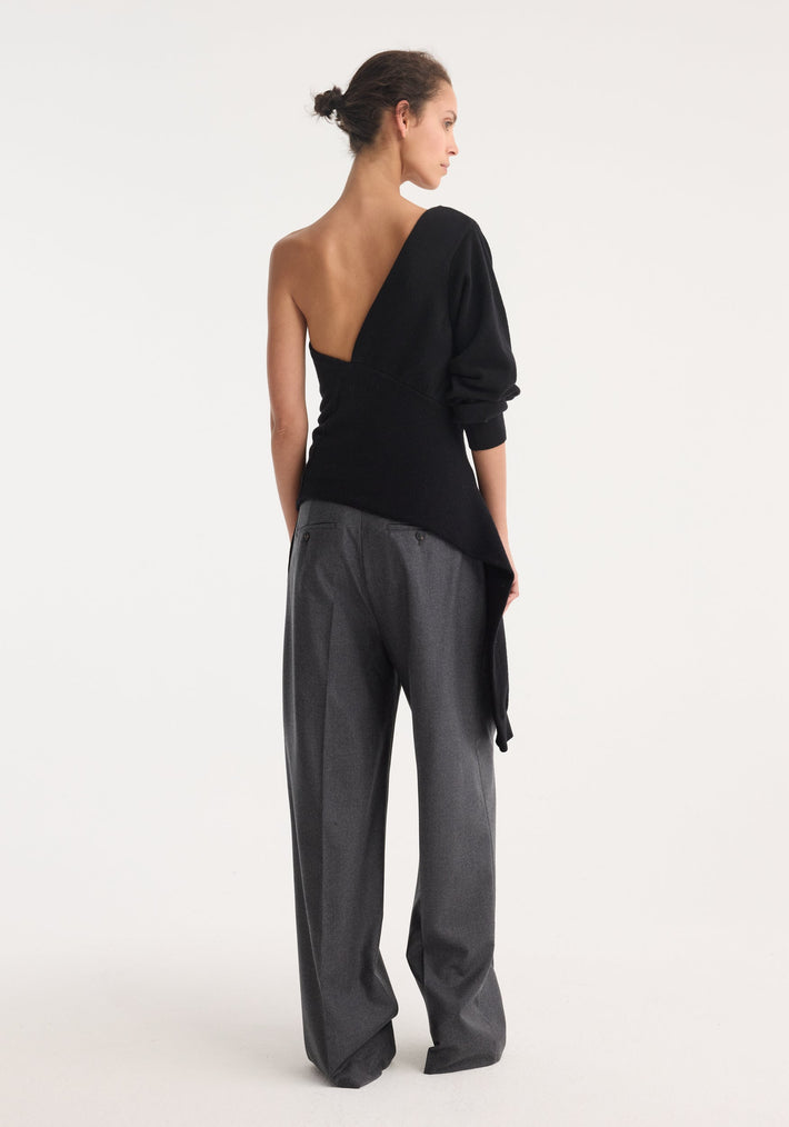 Model in an Off Shoulder Single Sleeve Top | Black, featuring an asymmetric design with one sleeve draped elegantly, paired with high-waisted, loose gray trousers.