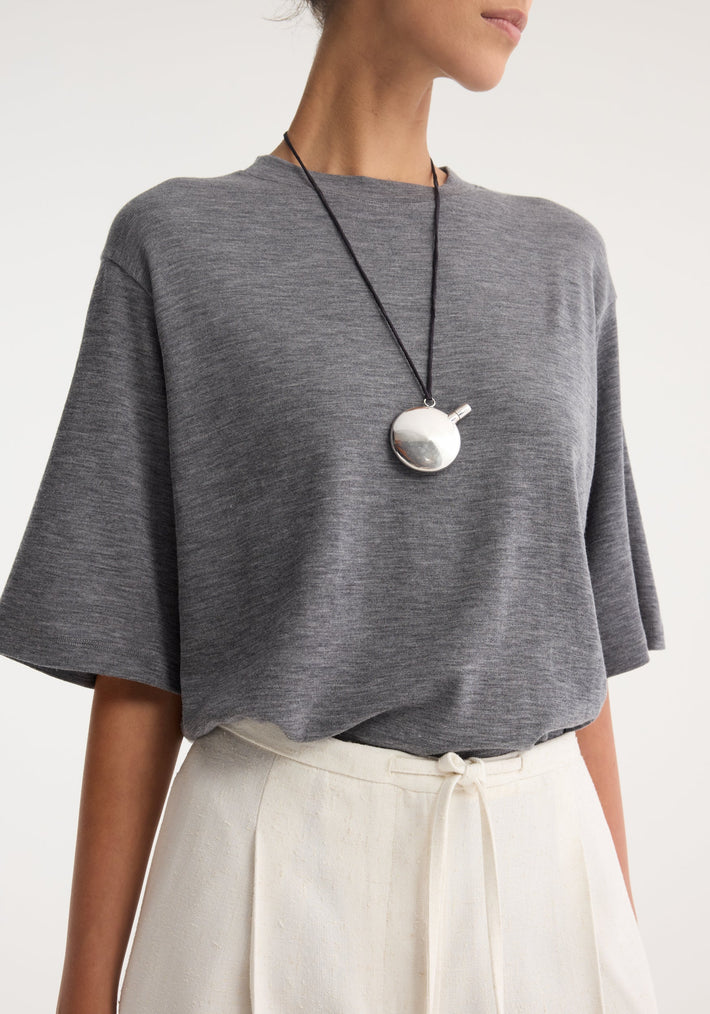 Person wearing a silver perfume bottle necklace on a black cord, featuring a polished pendant with spout detail, complementing a heather-gray top and off-white trousers.