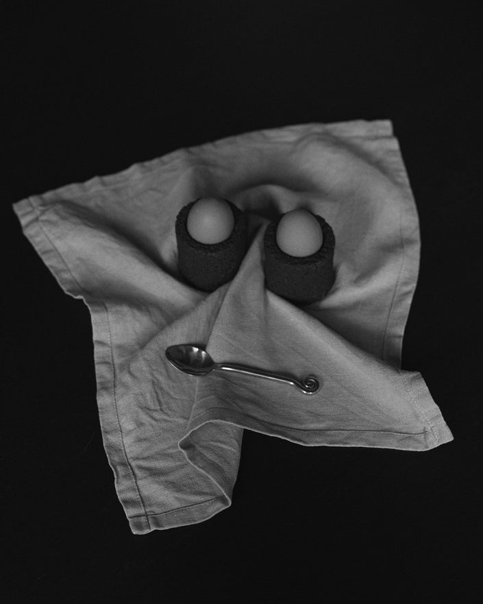 Egg Cups: Two handcrafted lava stone cups holding eggs, set on a linen napkin with a decorative teaspoon, showcasing unique craftsmanship by Dutch artist Louis Crombag.