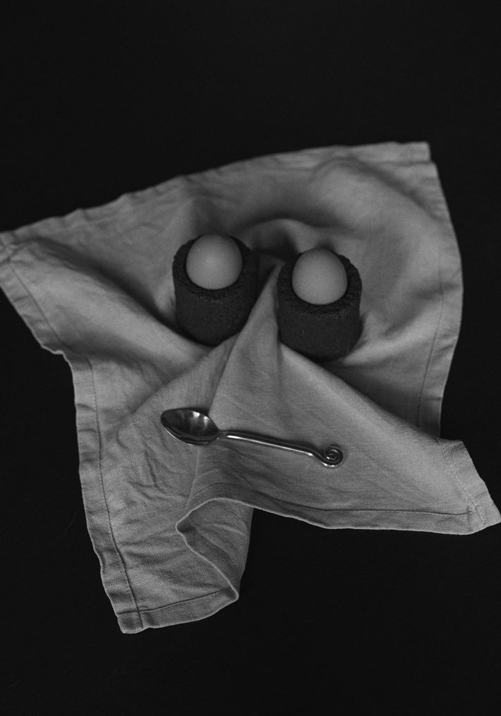 Egg Cups: Two handcrafted lava stone cups holding eggs, set on a linen napkin with a decorative teaspoon, showcasing unique craftsmanship by Dutch artist Louis Crombag.