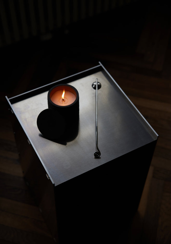 Candle Stopper | silver: A lit candle on a steel table accompanied by a spiraling stainless steel candle snuffer, elegantly designed for safe extinguishing.