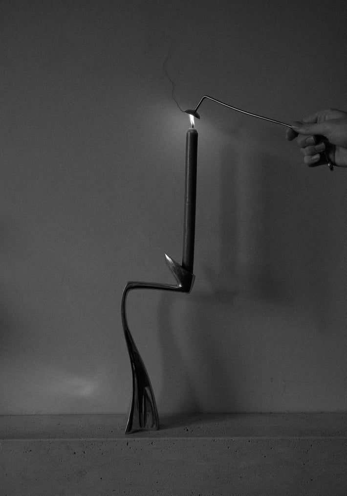 Silver Candle Stopper in a sculptural metal holder, twisting elegantly. A hand uses a tool to extinguish the flame, highlighting the holder's organic design.