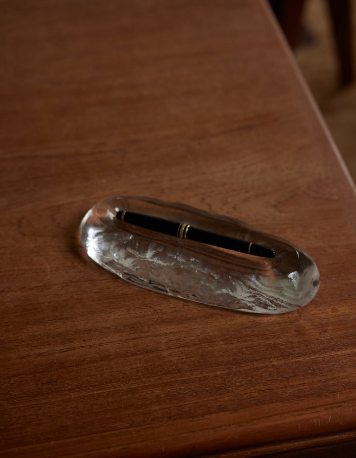 Close-up of Long trinket | Glass, an elegant clear glass pen rest, featuring a black pen with gold trim on a wooden desk.