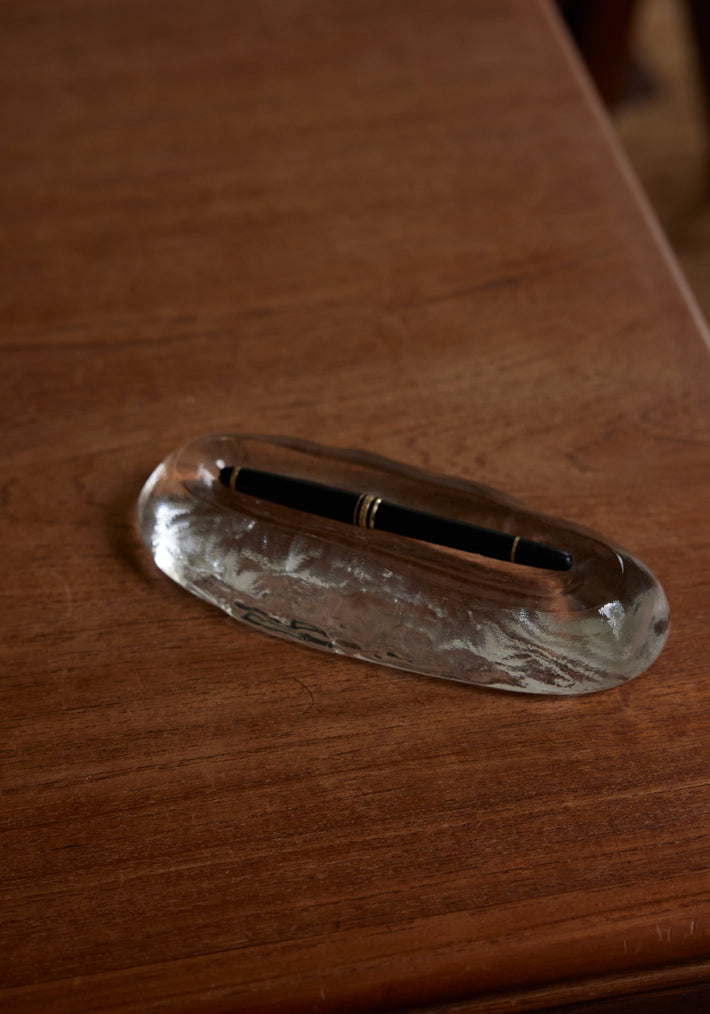 Close-up of Long trinket | Glass, an elegant clear glass pen rest, featuring a black pen with gold trim on a wooden desk.