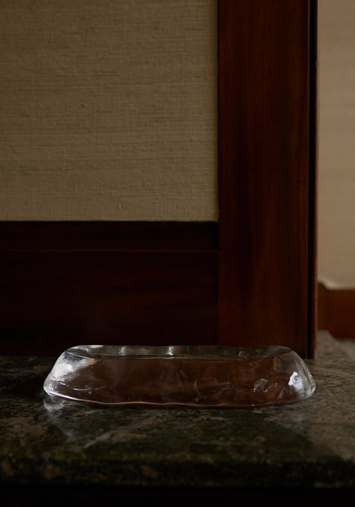 Long trinket | Glass: An irregularly shaped clear-glass dish by Szkło Studio, resting on a speckled stone surface, showcasing unique craftsmanship and experimental design.