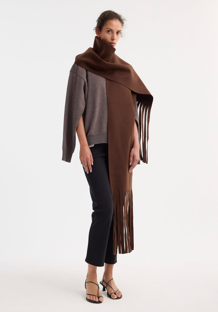 Woman wearing a long Wool scarf | pure chocolate, draped over a taupe sweater and black trousers, highlighting its fringed ends and luxurious virgin wool texture.