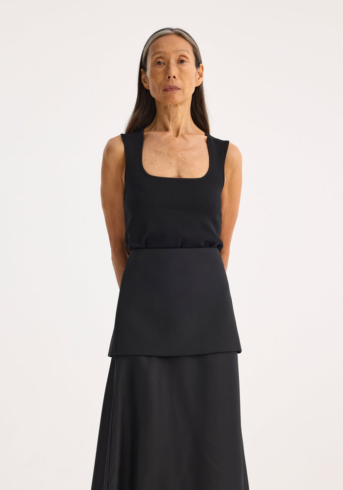 Woman wearing Bustier shaped knitted top with square neckline, fitted design, arms behind back, long skirt; minimalist fashion portrait.