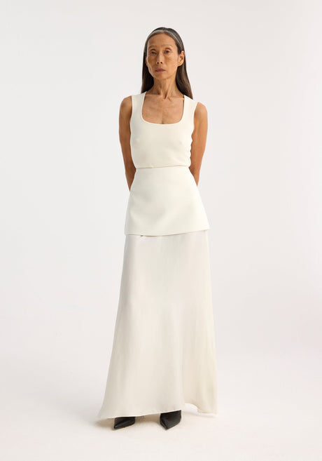 Person in a bustier-shaped knitted top, off white, with a square neckline, paired with a layered skirt. Arms behind back, neutral expression.