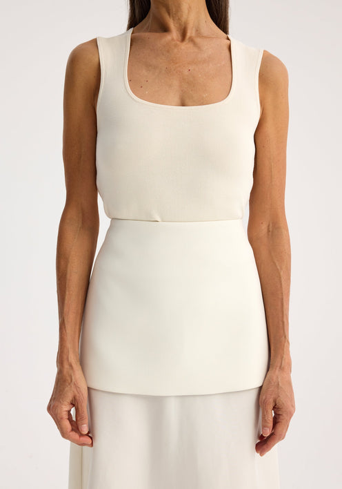 Person in a bustier-shaped knitted top, off white, with a square neckline, paired with a layered skirt. Arms behind back, neutral expression.