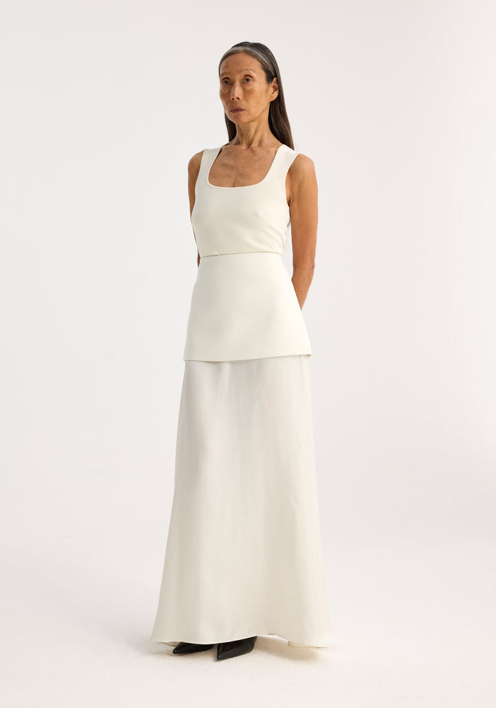 Bustier shaped knitted top in off white, featuring a scoop neck and peplum, worn by a slim, mature model with a neutral expression.