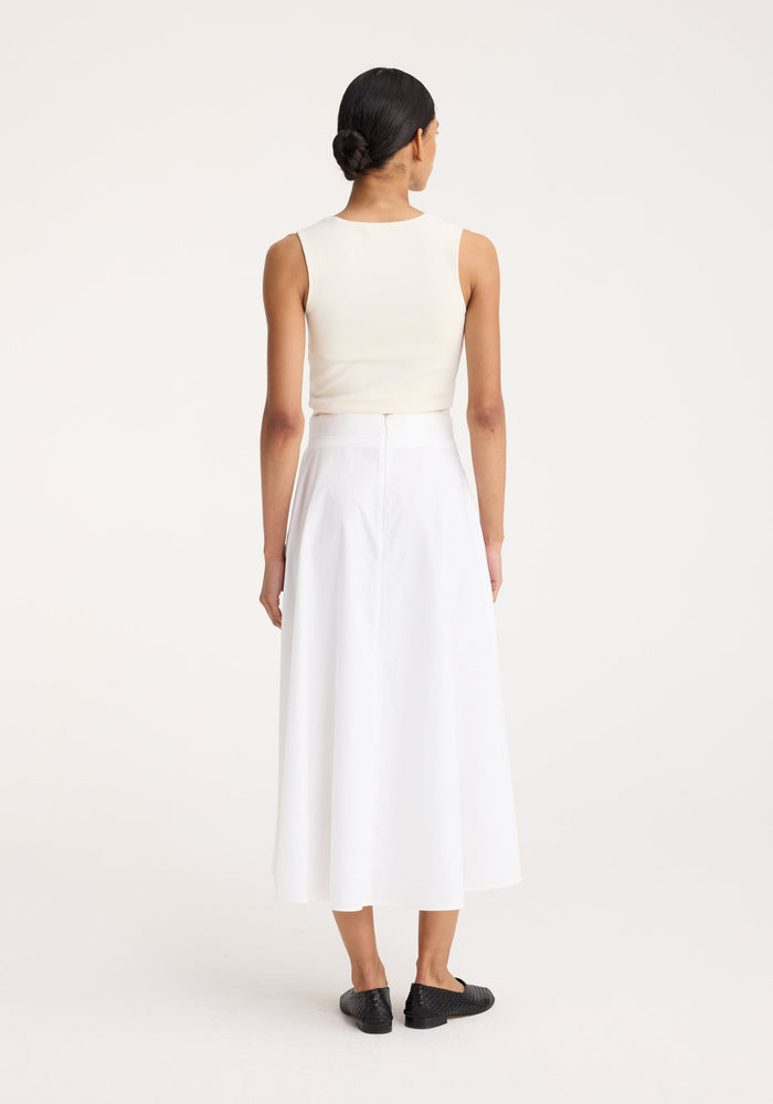 Woman in off-white bustier-shaped knitted top with fitted design, viewed from the back, paired with a high-waisted white skirt and black loafers.