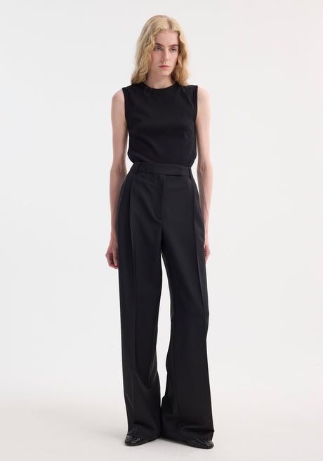 Slim individual wearing a Dense rib tank top in black, fitted and sleeveless, with high-waisted wide-leg trousers, showcasing minimalist fashion in a studio setting.