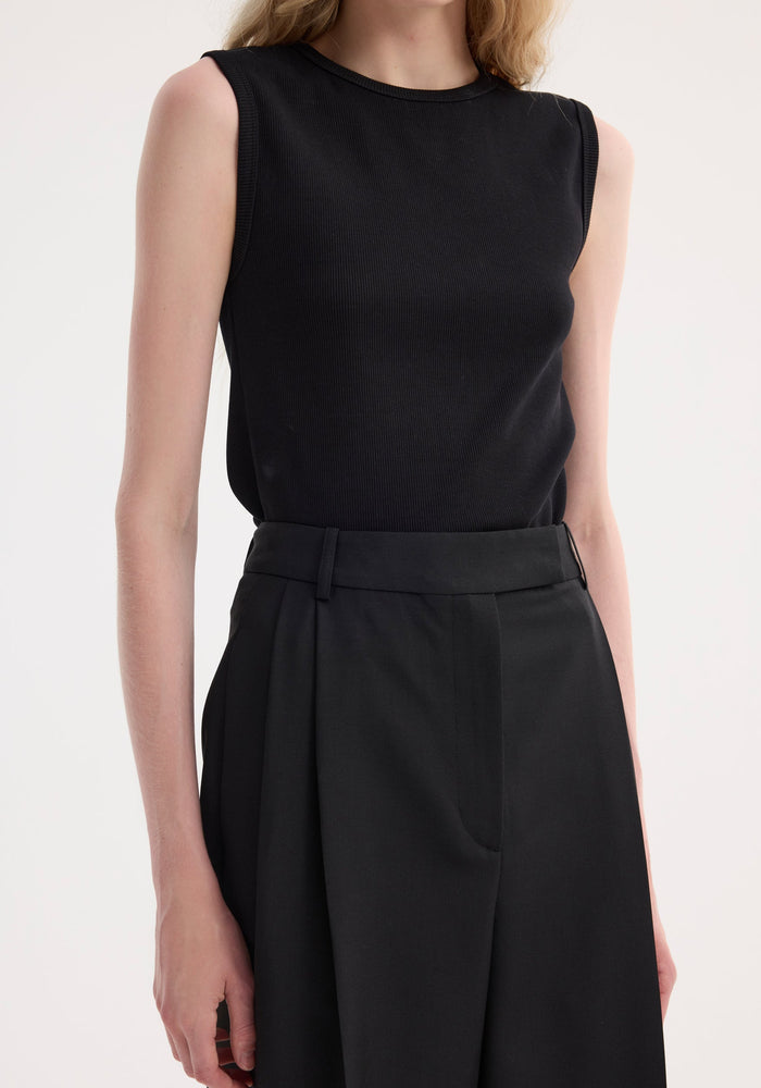 Person wearing a Dense rib tank top in black, crafted from organic cotton and elastane, tucked into high-waisted trousers, showcasing its fitted design.