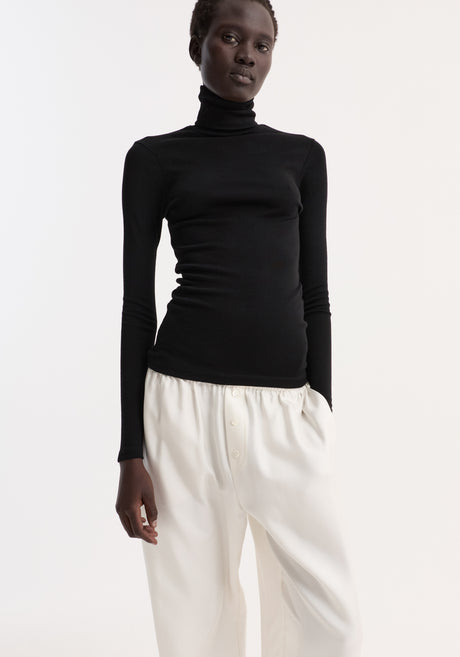 Fine merino turtleneck in black, worn by a model with loose high-waisted trousers and flat sandals, showcasing a minimalist, streamlined look.