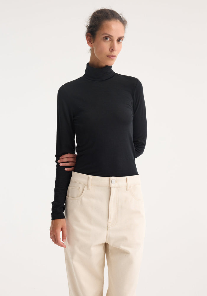 Fine merino turtleneck | black, worn by a model with tucked-in high-waisted trousers, showcasing its fitted silhouette and long sleeves. Ideal for layering and versatile styling.