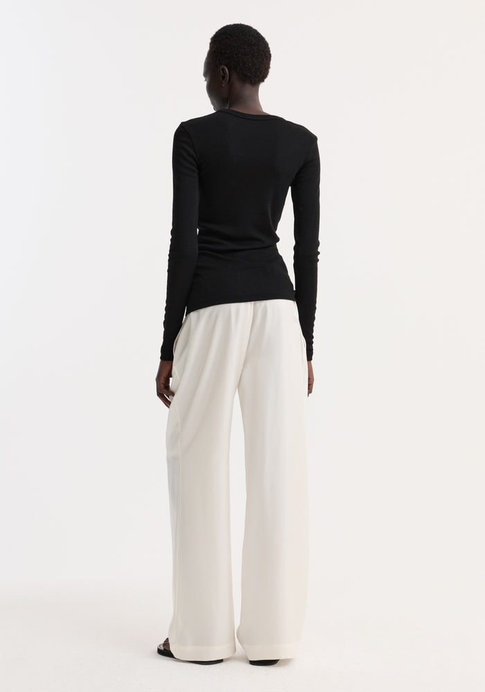 Fine merino long sleeve | black: Model wears fitted black merino wool top, crew neck, long sleeves, paired with high-waisted white trousers, highlighting minimalist elegance.