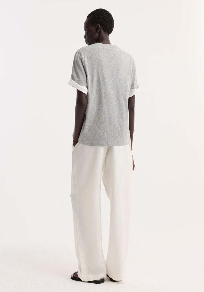 Person wearing Slub melange cotton t-shirt in light grey, oversized fit, crewneck, with rolled sleeves, paired with loose white trousers and black sandals, seen from behind.