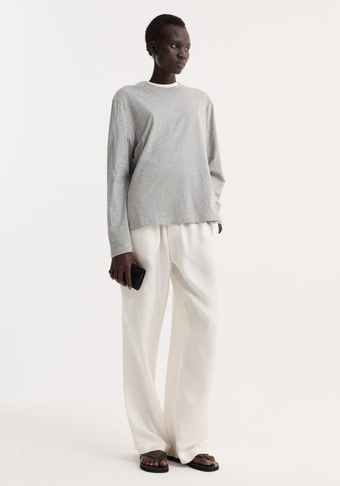 Woman wearing Slub melange cotton long sleeve in light grey, tucked into high-waisted white trousers, holding a black wallet, styled with black sandals.
