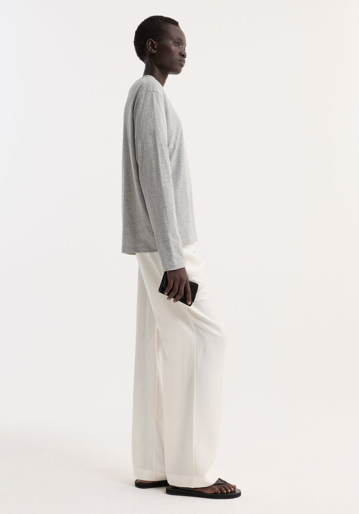 Person in profile wearing a Slub melange cotton long sleeve in light grey melange, paired with loose white trousers and black flat slide sandals, holding a black wallet.