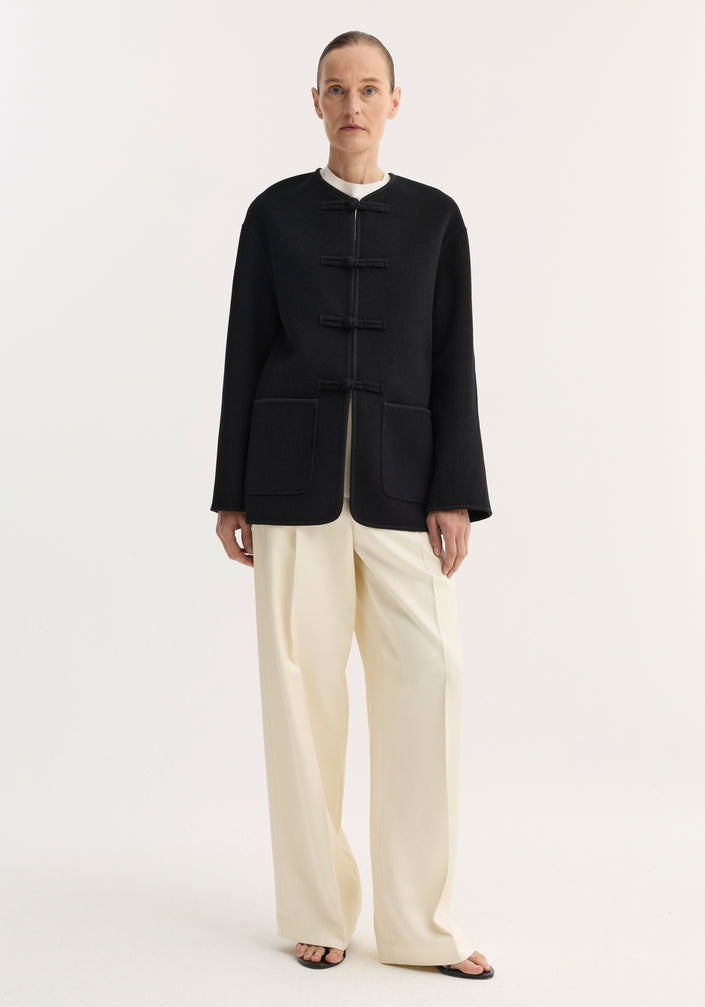 Model wearing the Mandarin double-faced wool jacket in black, featuring frog-style closures, embroidered edges, and patch pockets, paired with wide-leg trousers and slide sandals.