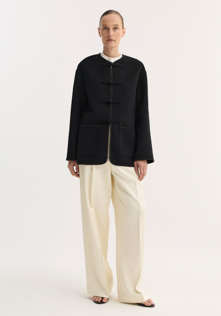 Model wearing the Mandarin double-faced wool jacket in black, featuring frog-style closures, embroidered edges, and patch pockets, paired with wide-leg trousers and slide sandals.