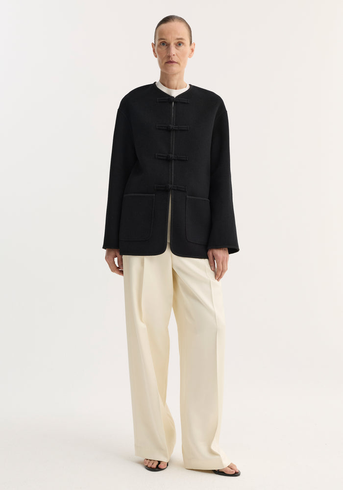 Model wearing double-faced wool pankou jacket in black, featuring traditional frog toggle closures, embroidered edges, and patch pockets, paired with cream trousers and sandals.