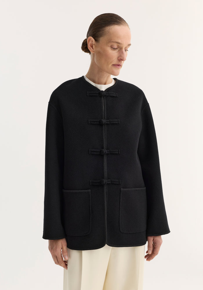 Model in black double-faced wool pankou jacket with frog-style toggles, embroidered edges, and large patch pockets, paired with cream trousers.