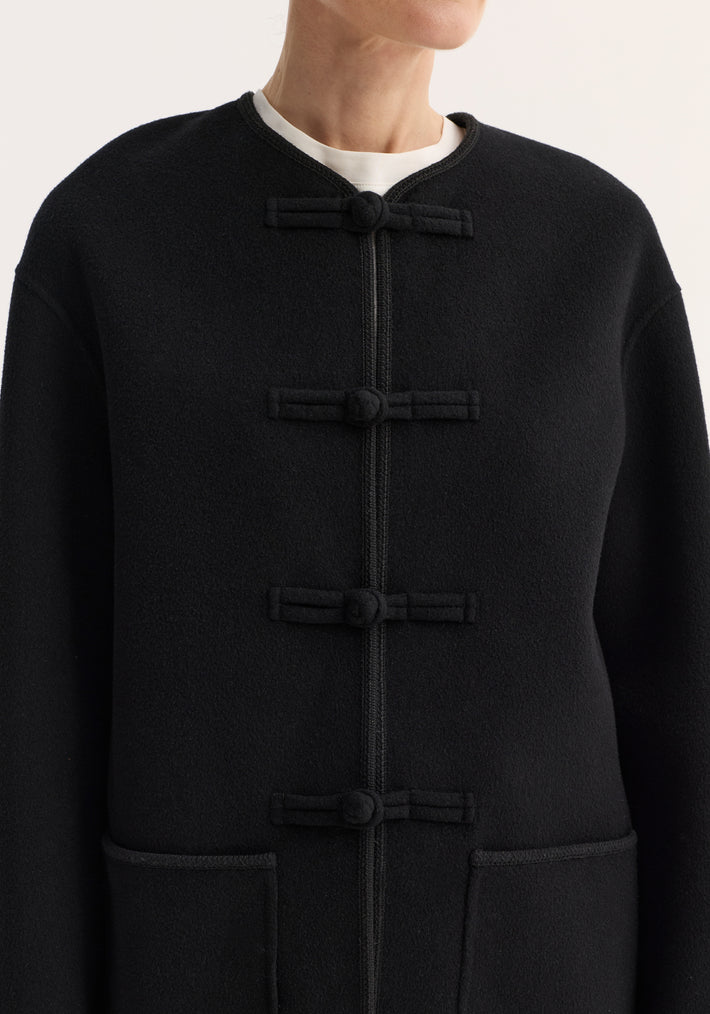 Close-up of a double-faced wool pankou jacket with traditional frog closures, embroidered edges, patch pockets, and a round neckline, worn over a visible white shirt.