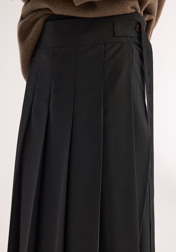 Close-up of Plissé wrap skirt in black, featuring crisp pleats, tailored waistband with button tab, and brown sweater tucked in, showcasing its refined silhouette.