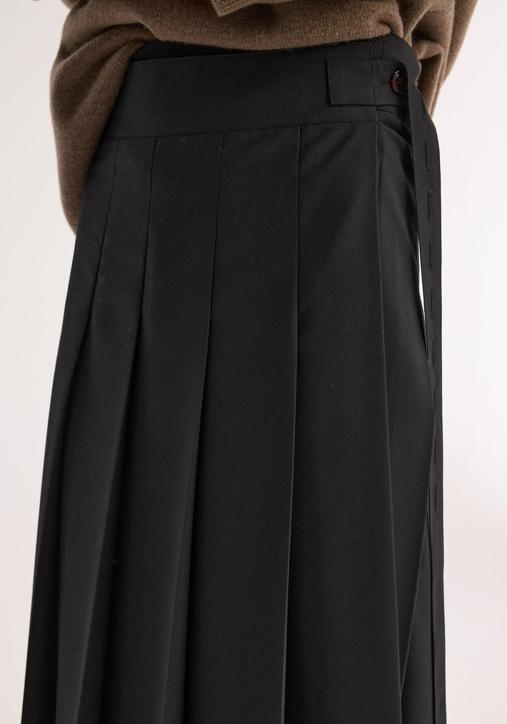 Close-up of Plissé wrap skirt in black, featuring crisp pleats, tailored waistband with button tab, and brown sweater tucked in, showcasing its refined silhouette.