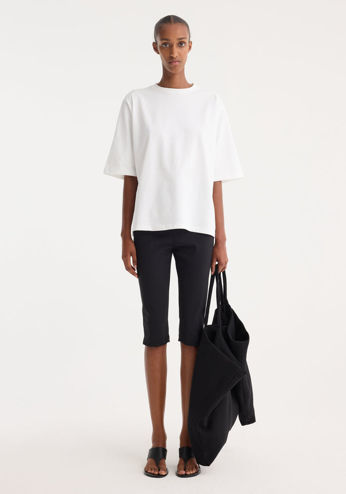 Woman wearing Scuba capri | black, showcasing tailored, knee-length scuba fabric trousers with a structured fit, styled with a loose white T-shirt and black sandals.