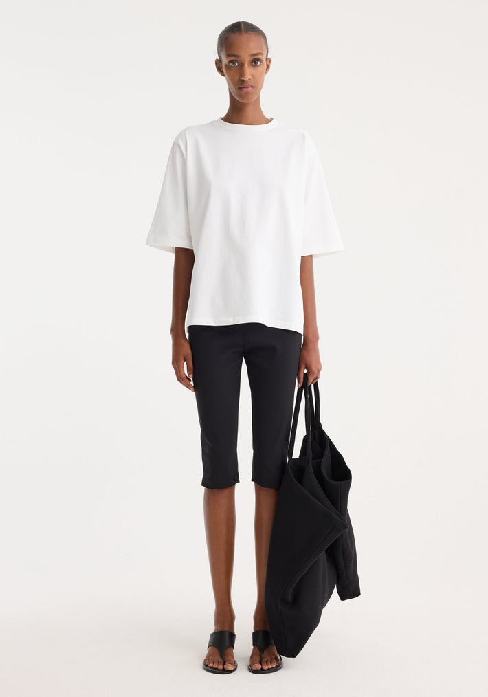 Woman wearing Scuba capri | black, showcasing tailored, knee-length scuba fabric trousers with a structured fit, styled with a loose white T-shirt and black sandals.