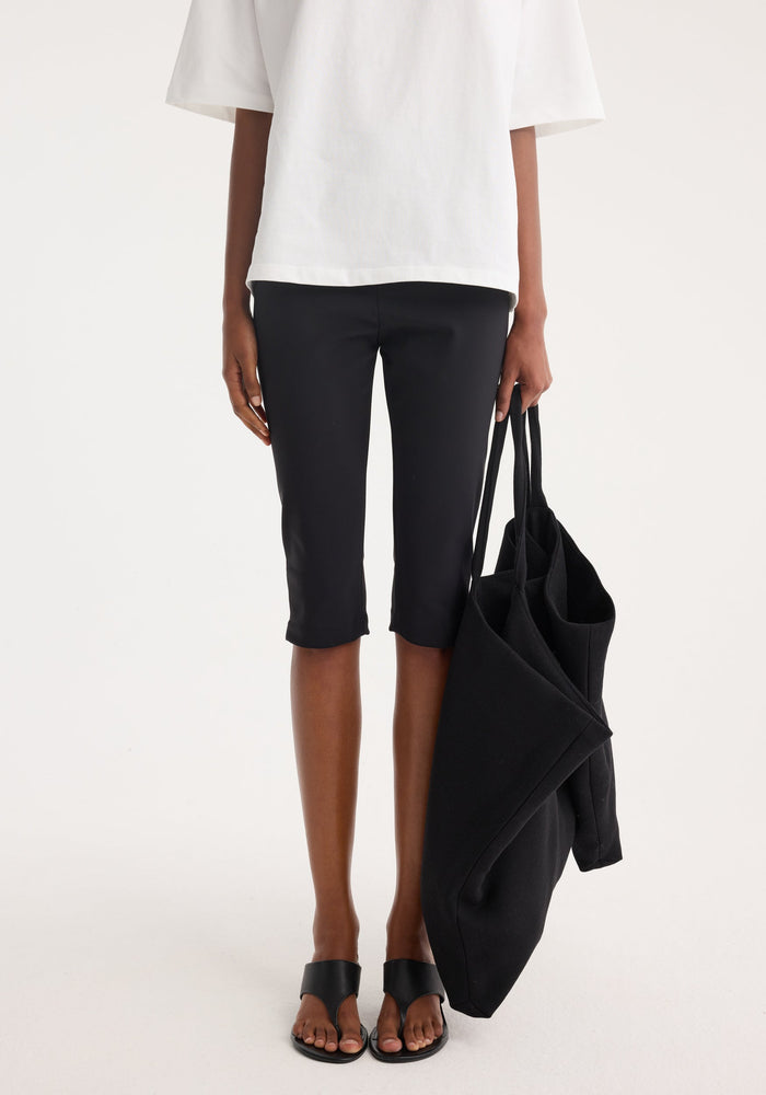 Model wearing Scuba capri | black, showcasing slim, knee-length fit in heavyweight scuba fabric, paired with black sandals and holding a large black tote bag.