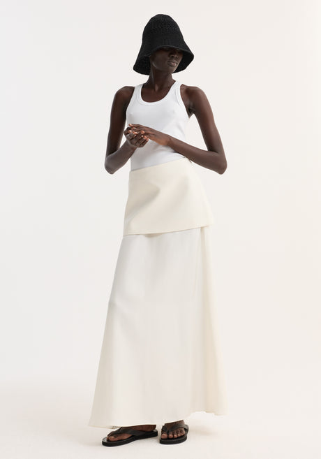 Model wearing Integrated fluid silk skirt, cream, with asymmetrical overlay and high waist, paired with white tank, black bucket hat, and black sandals.
