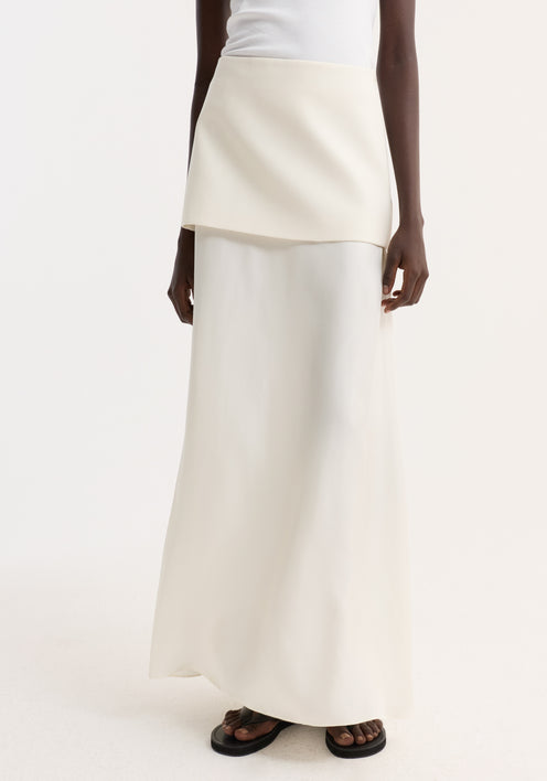 Model wearing Integrated fluid silk skirt, cream, with asymmetrical overlay and high waist, paired with white tank, black bucket hat, and black sandals.