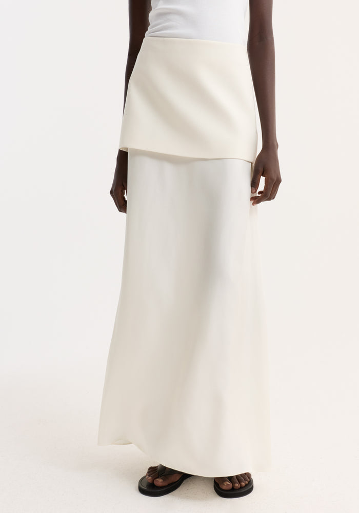 Integrated fluid silk skirt in cream, featuring a high-waisted, floor-length design with a sculptural silhouette, worn with black sandals against a neutral backdrop.