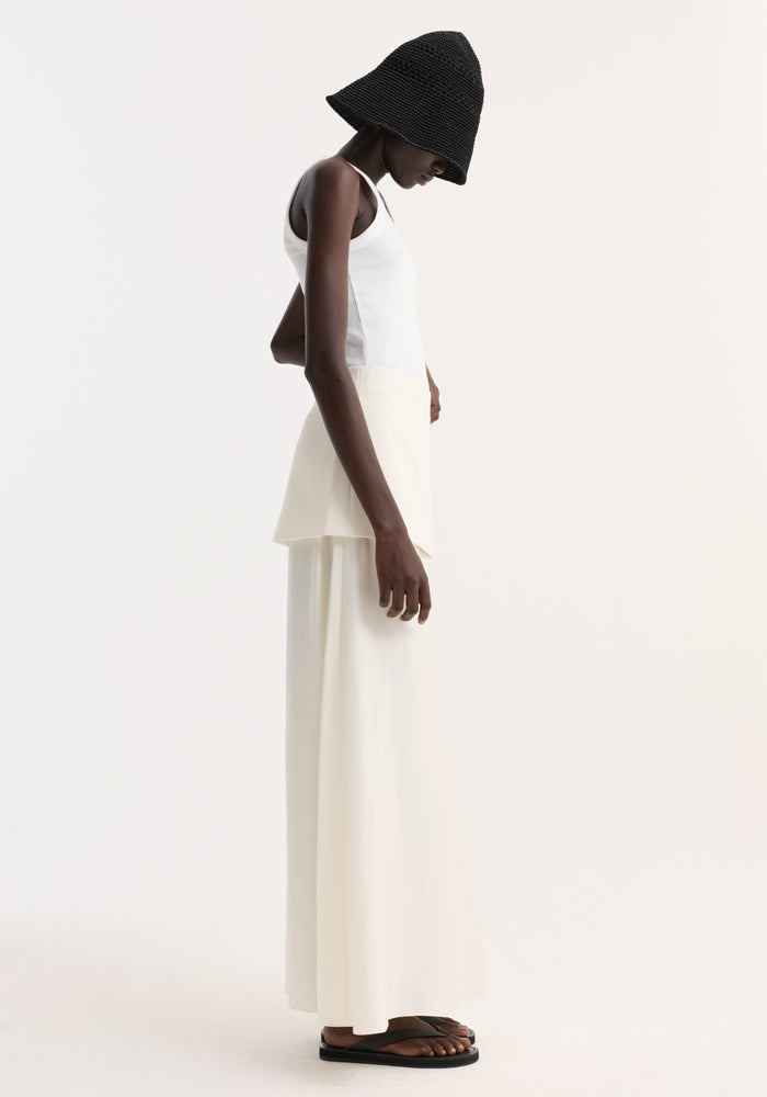 Model in an Integrated fluid silk skirt | cream, showcasing its high waist, layered panel detail, and flowing silhouette, paired with a white tank and black bucket hat.