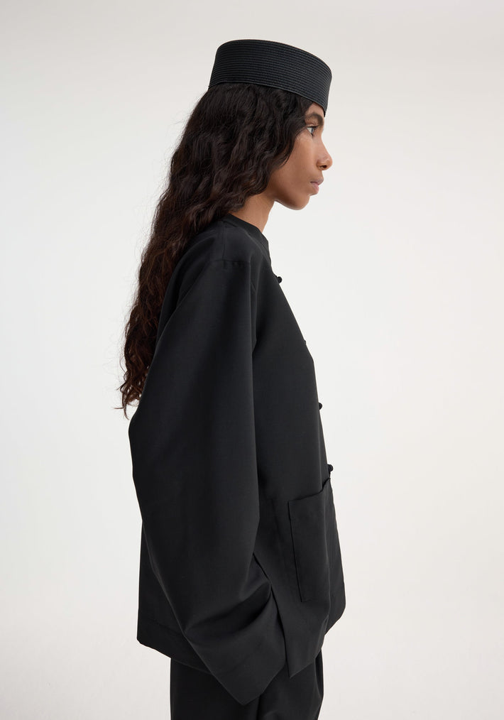 Side-profile of a woman wearing the Mandarin jacket | black, featuring a collarless design, pankou closures, and patch pockets, paired with matching trousers and a pillbox hat.