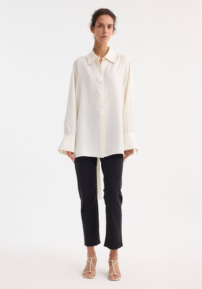 Model wearing a Deconstructed silk shirt in cream, featuring oversized fit, extended cuffs, open back, and versatile tie detail, paired with black trousers and white sandals.