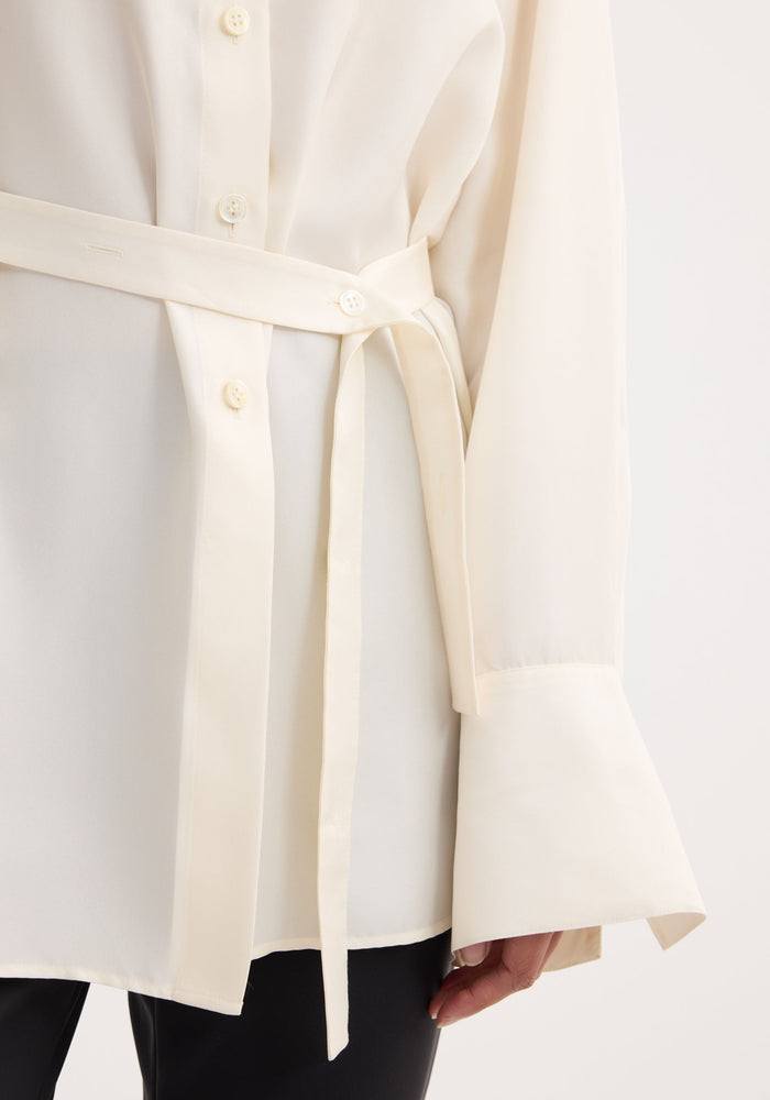 Deconstructed silk shirt in cream with wide cuffs and self-fabric belt, featuring an open back and versatile tie detail. Worn with black trousers.