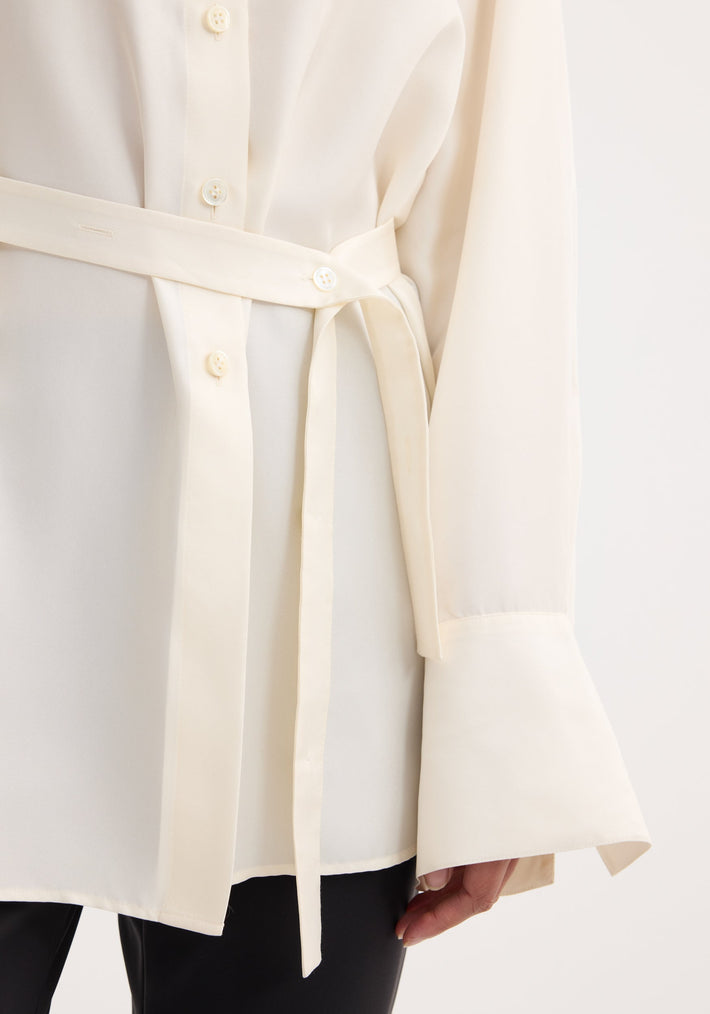 Deconstructed silk shirt in cream with wide cuffs and self-fabric belt, featuring an open back and versatile tie detail. Worn with black trousers.
