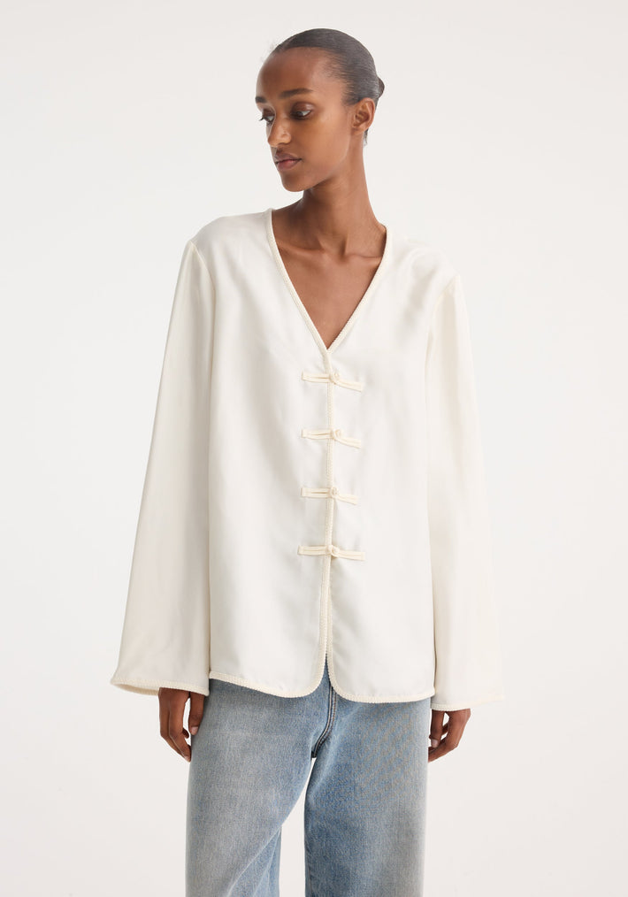Model wearing Mandarin silk top in cream with a V-neck, pankou closures, embroidered edges, and long sleeves, paired with straight-leg jeans.