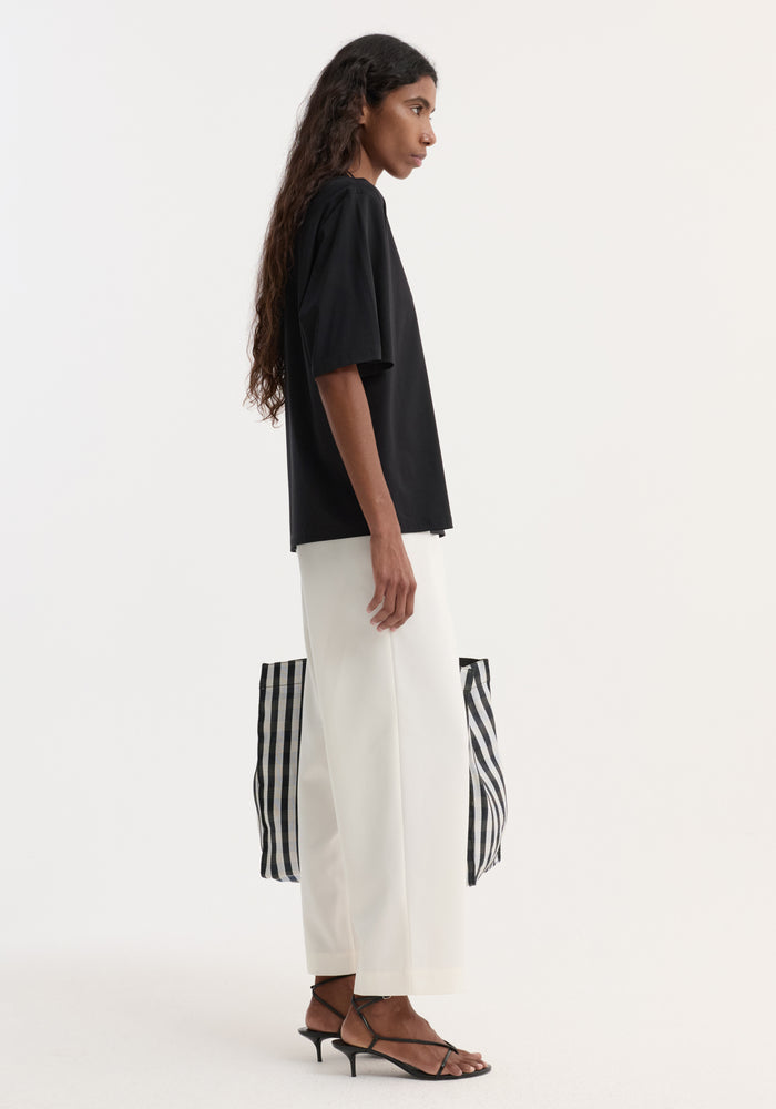 Side-profile of a person wearing an Oversized cotton t-shirt with a crew neck and dropped shoulders, paired with white trousers and carrying a checked tote bag.