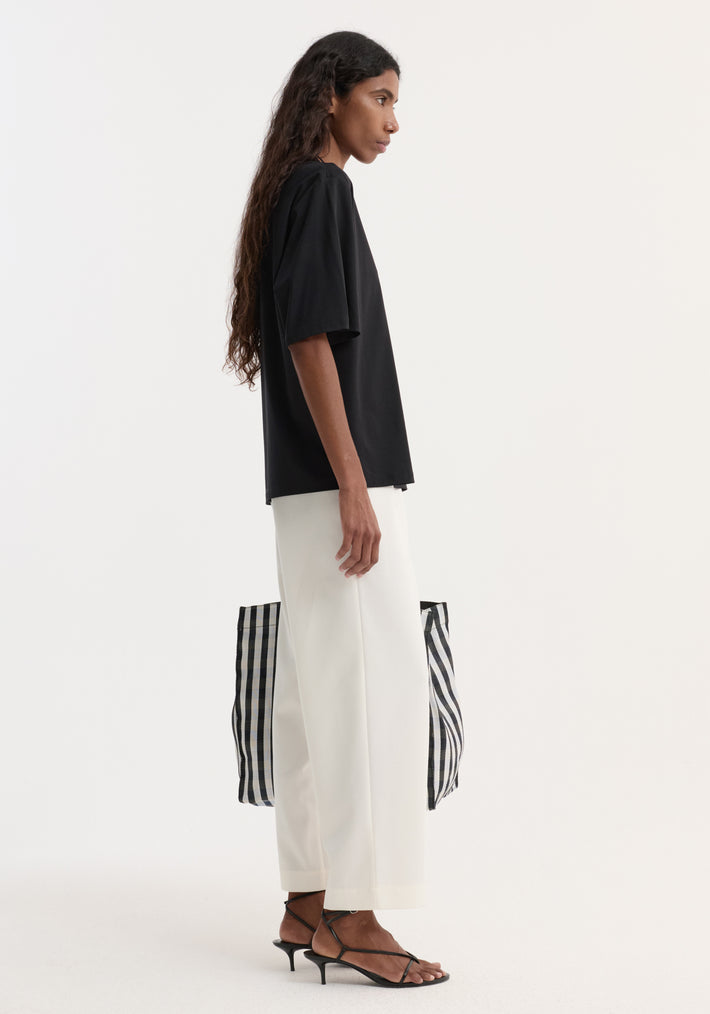 Side-profile of a person wearing an Oversized cotton t-shirt with a crew neck and dropped shoulders, paired with white trousers and carrying a checked tote bag.